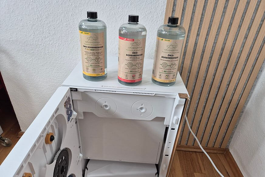 Grashüpfer Produktübersicht aller Öko-Reiniger für Wischroboter stehen auf der Wischroboter Service-Station nebeneinander, Zitrusblüte, Tropisch und Pink-Grapefruit